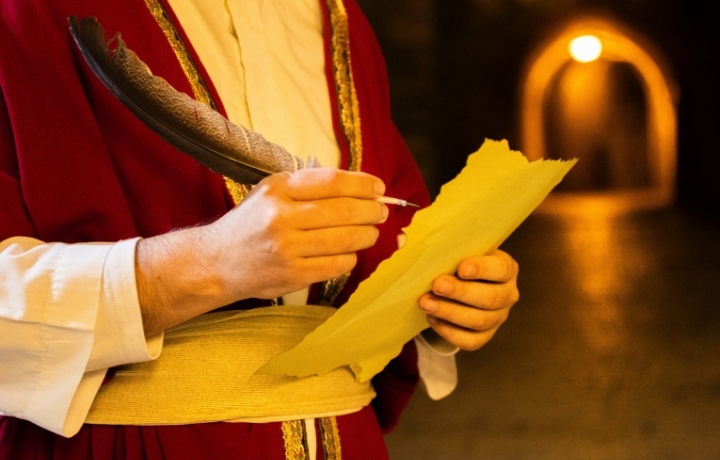 Mann in mittelalterlichem Gewand schreibt mit Feder auf Papier.