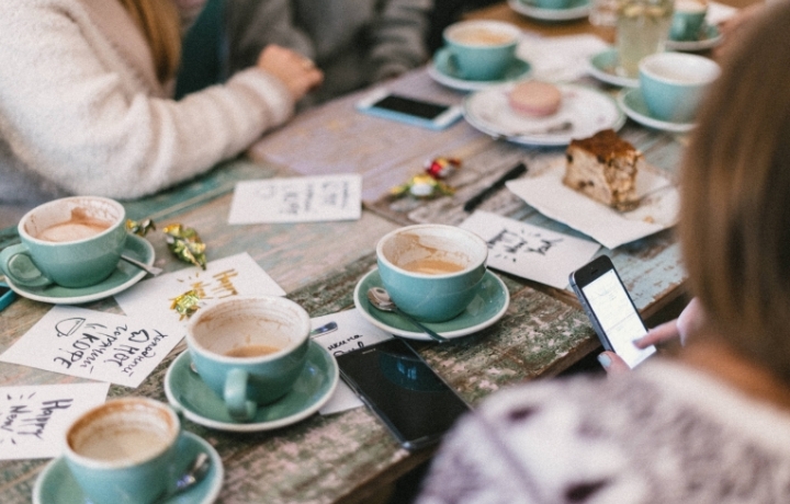 Viele türkisfarbene Kaffeetassen auf einem Holztisch, zwei Frauen schemenhaft zu erkennen, mit Handys in der Hand.