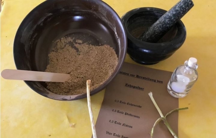 Selbsthergestelltes Zahnpulver in einer schwarzen Schüssel mit Spatel drin. Gelber Tisch.